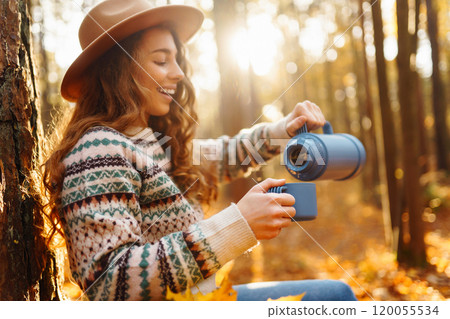 Cute young woman drinks hot drink from thermos and enjoys nature. Autumn landscape. relax concept. 120055534