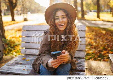 Young smiling woman sitting on bench in autumn park using phone in stylish clothes happy mood. Young smiling woman sitting on bench in autumn park using phone in stylish clothes happy mood. 120055572