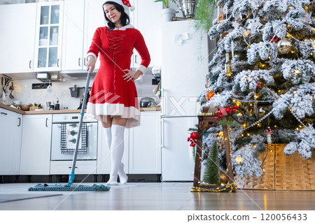 A woman in a red dress and Santa hat cleans the floor with a mop in a white kitchen with Christmas decor and a Christmas tree. Cleaning the house and preparing for the holidays Christmas and New Year A woman in a red dress and Santa hat cleans the floor with a mop in a white kitchen with Christmas decor and a Christmas tree. Cleaning the house and preparing for the holidays Christmas and New Year 120056433