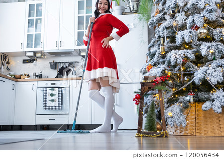 A woman in a red dress and Santa hat cleans the floor with a mop in a white kitchen with Christmas decor and a Christmas tree. Cleaning the house and preparing for the holidays Christmas and New Year A woman in a red dress and Santa hat cleans the floor with a mop in a white kitchen with Christmas decor and a Christmas tree. Cleaning the house and preparing for the holidays Christmas and New Year 120056434