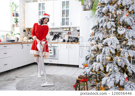 A woman in a red dress and Santa hat cleans the floor with a vacuum cleaner in white kitchen with Christmas decor. Cleaning the house and preparing for the holidays Christmas and New Year A woman in a red dress and Santa hat cleans the floor with a vacuum cleaner in white kitchen with Christmas decor. Cleaning the house and preparing for the holidays Christmas and New Year 120056446
