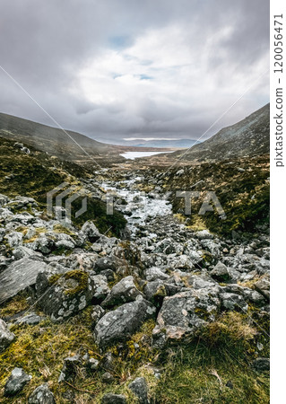 A rocky, grassy valley with a small stream flowing through it. The hills on either side are covered in vegetation, and the sky is cloudy. A rocky, grassy valley with a small stream flowing through it. The hills on either side are covered in vegetation, and the sky is cloudy. 120056471
