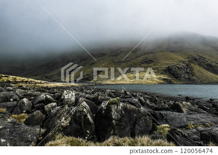 A large body of water sits nestled at the base of a misty, green-covered mountain. The water is calm, reflecting the overcast sky. Scattered rocks line the shoreline. 120056474