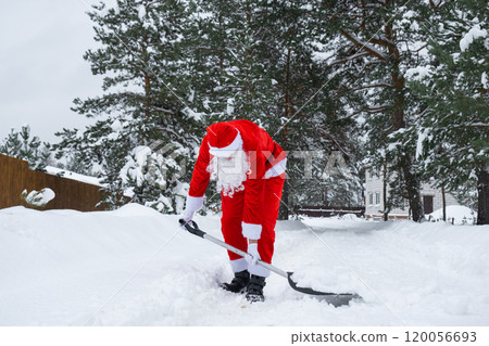 Santa Claus cleans snow with shovel in winter outdoors after a snowfall. Cleaning the streets in the village, clearing the passage for cars, difficult weather conditions for Christmas and New Year Santa Claus cleans snow with shovel in winter outdoors after a snowfall. Cleaning the streets in the village, clearing the passage for cars, difficult weather conditions for Christmas and New Year 120056693