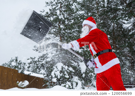 Santa Claus cleans snow with shovel in winter outdoors after a snowfall. Cleaning the streets in the village, clearing the passage for cars, difficult weather conditions for Christmas and New Year 120056699