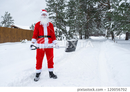 Santa Claus cleans snow with shovel in winter outdoors after a snowfall. Cleaning the streets in the village, clearing the passage for cars, difficult weather conditions for Christmas and New Year Santa Claus cleans snow with shovel in winter outdoors after a snowfall. Cleaning the streets in the village, clearing the passage for cars, difficult weather conditions for Christmas and New Year 120056702