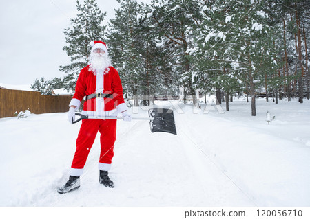 Santa Claus cleans snow with shovel in winter outdoors after a snowfall. Cleaning the streets in the village, clearing the passage for cars, difficult weather conditions for Christmas and New Year 120056710