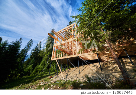 Residential building with wooden frame in process of being built near a forest. Beginning of new construction of comfortable cottage. Idea of ecological construction, contemporary architectural design Residential building with wooden frame in process of being built near a forest. Beginning of new construction of comfortable cottage. Idea of ecological construction, contemporary architectural design 120057445