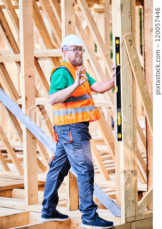 Carpenter constructing wooden frame house. Man inspects walls for levelness using spirit level, showing gesture OK sign, wearing protective overalls, helmet and orange vest. Carpenter constructing wooden frame house. Man inspects walls for levelness using spirit level, showing gesture OK sign, wearing protective overalls, helmet and orange vest. 120057446