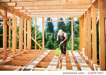 Laborer constructing wooden frame house near forest. Man treating woods, applying fire retardant using sprayer, while dressed in protective suit, helmet. Concept of modern eco-friendly construction. 120057447