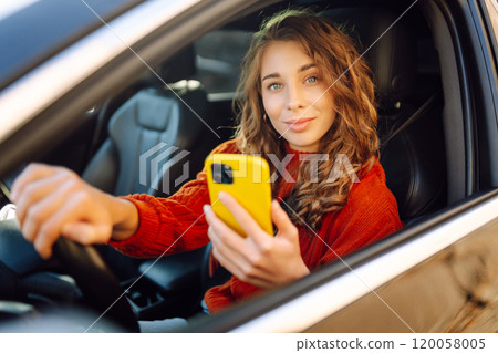 Portrait of a young woman sitting in a car in the driver's seat looking into a smartphone. 120058005