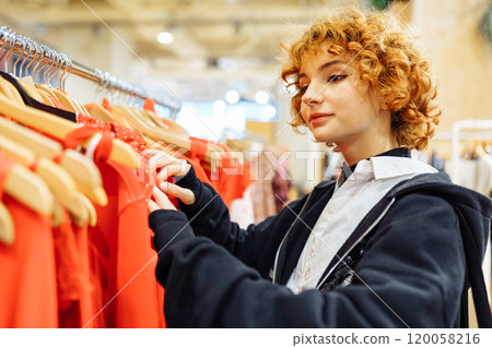 young attractive red-haired woman choosing red clothes in boutique young attractive red-haired woman choosing red clothes in boutique 120058216