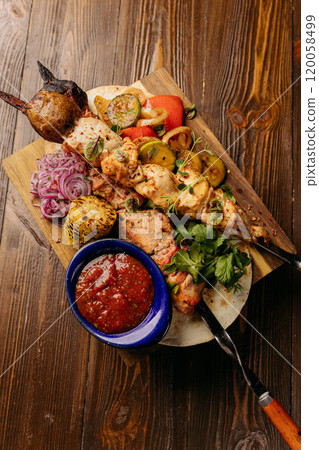 Top view of grilled skewers with vegetables, served on a wooden board with spicy dipping sauce 120058499