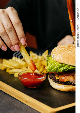 Dipping crispy French fry into ketchup beside a juicy burger with fresh lettuce, food moment 120058508