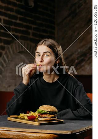 Playful woman enjoys a crispy fry with burger and fries meal in a cozy restaurant setting Playful woman enjoys a crispy fry with burger and fries meal in a cozy restaurant setting 120058509