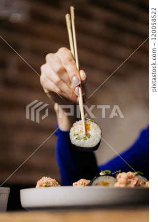 Hand holding sushi roll with chopsticks, showcasing fresh ingredients of cucumber and crab Hand holding sushi roll with chopsticks, showcasing fresh ingredients of cucumber and crab 120058522