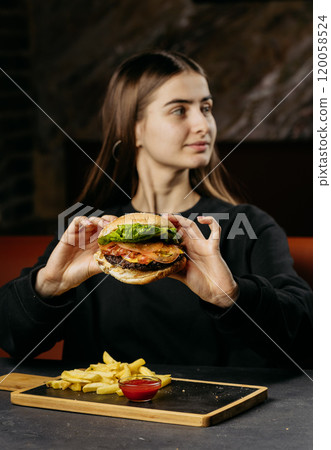 Woman holding a juicy burger with fries on the side, looking away in a casual restaurant setting Woman holding a juicy burger with fries on the side, looking away in a casual restaurant setting 120058524