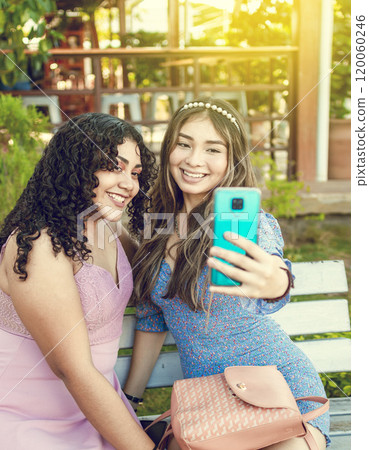 Two pretty girls sitting on a bench taking a selfie, girls smiling and taking a selfie, sisterly friendship concept 120060246
