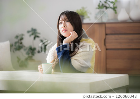 A young woman relaxing in the living room in winter drinking a hot drink; She has too much to do and is resting her chin on her hand (putting her chin on her hand), worried and troubled 120060348