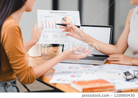 Two professionals analyze business data using laptop displaying sales dashboard and various printed charts on desk. 120060430