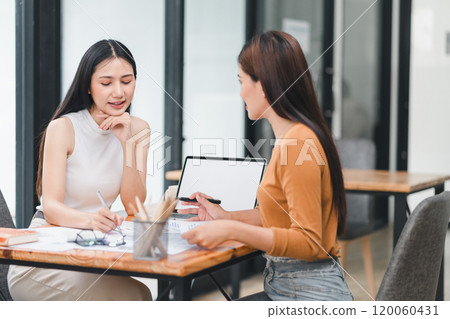 Two young Asian businesswomen discuss a new startup project idea, analyzing planning and financial statistics, investment market, at the office. Two young Asian businesswomen discuss a new startup project idea, analyzing planning and financial statistics, investment market, at the office. 120060431