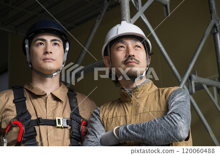 The serious and earnest frontal expressions of two construction workers and blue collar workers working outdoors wearing safety harnesses looking up The serious and earnest frontal expressions of two construction workers and blue collar workers working outdoors wearing safety harnesses looking up 120060488