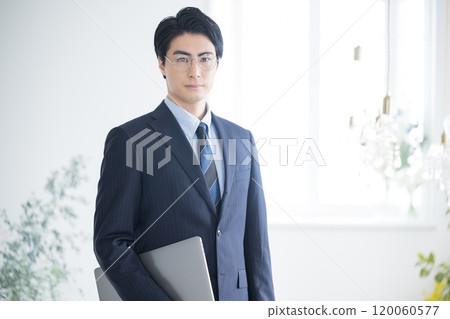 A young businessman looking at the camera while holding a computer, such as a tax accountant, accountant, or consultant A young businessman looking at the camera while holding a computer, such as a tax accountant, accountant, or consultant 120060577