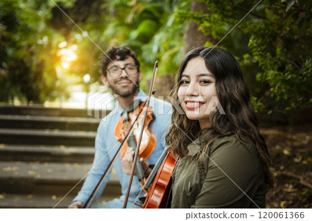 Two young violinists sitting outdoors looking at the camera. Portrait of two young violists sitting outside. Man and woman violinist sitting on the stairs. Concept of two young violinists 120061366