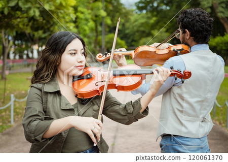 Two young violinists standing playing violin in a park. Portrait of man and woman together playing violin in park. Violinist man and woman back to back playing violin in a park outdoors Two young violinists standing playing violin in a park. Portrait of man and woman together playing violin in park. Violinist man and woman back to back playing violin in a park outdoors 120061370