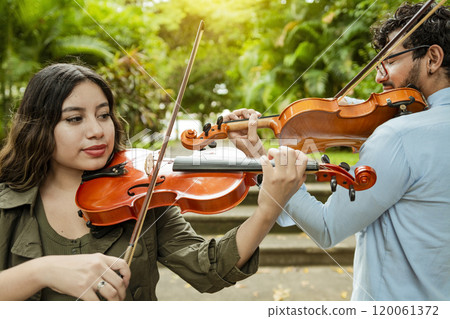 Violinist man and woman back to back playing violin in a park outdoors. Two young violinists standing playing violin in a park. Portrait of man and woman together playing violin in park Violinist man and woman back to back playing violin in a park outdoors. Two young violinists standing playing violin in a park. Portrait of man and woman together playing violin in park 120061372