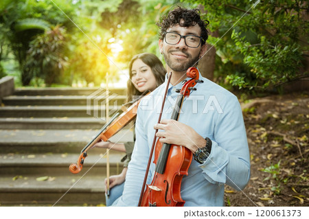 Portrait of two young violists sitting outside. Man and woman violinist sitting on the stairs. Concept of two young violinists, Two young violinists sitting outdoors looking at the camera Portrait of two young violists sitting outside. Man and woman violinist sitting on the stairs. Concept of two young violinists, Two young violinists sitting outdoors looking at the camera 120061373