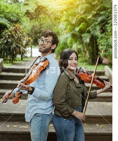 Portrait of two young violists back to back outdoors. Portrait of smiling male and female violinist back to back outdoors. Two young violinists back to back holding their violins Portrait of two young violists back to back outdoors. Portrait of smiling male and female violinist back to back outdoors. Two young violinists back to back holding their violins 120061374