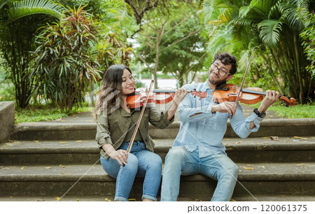 Portrait of two young violists playing violin sitting on stairs outside. Portrait of smiling violinists sitting on stairs playing violin. Man and woman sitting on stairs playing violin 120061375