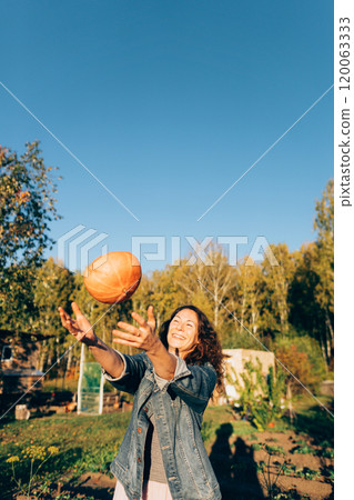 Joyful woman tossing a pumpkin on a sunny autumn day in the countryside. Middle-aged woman celebrating the harvest season in her garden with a pumpkin 120063333