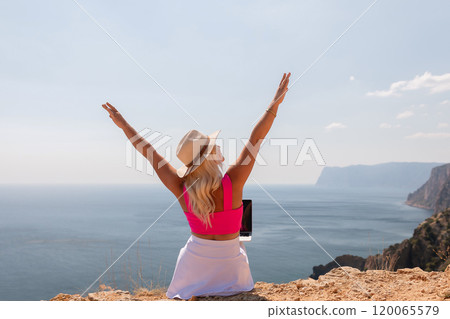 A woman is sitting on a hill overlooking the ocean with a laptop in her lap. She is wearing a pink top and a straw hat. Concept of relaxation and leisure, as the woman enjoys the view. A woman is sitting on a hill overlooking the ocean with a laptop in her lap. She is wearing a pink top and a straw hat. Concept of relaxation and leisure, as the woman enjoys the view. 120065579
