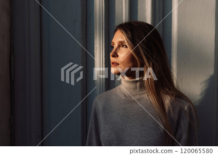 A woman with long hair is standing in front of a white door. She is wearing a sweater and she is looking out the window. 120065580