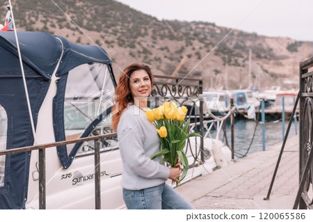 Woman holds yellow tulips in harbor with boats docked in the background., overcast day, yellow sweater, mountains Woman holds yellow tulips in harbor with boats docked in the background., overcast day, yellow sweater, mountains 120065586