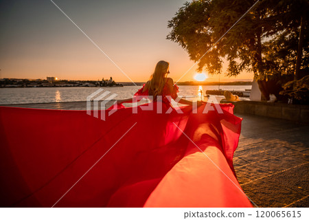 Sunrise red dress. A woman in a long red dress against the backdrop of sunrise, bright golden light of the sun's rays. The concept of femininity, harmony. 120065615