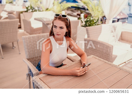 Woman sits in outdoor cafe at wooden table, holds smartphone. Bright sunny day provides natural lighting. Cafe offers relaxed atmosphere for customers to enjoy refreshments. 120065631