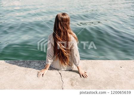 A woman is sitting on a ledge by a body of water, wearing a dress with a leopard print pattern. The scene is peaceful and serene, with the woman enjoying the view of the water and the surrounding area 120065639
