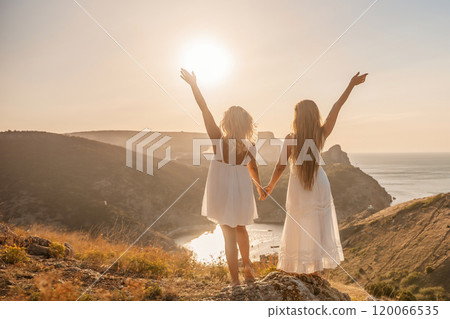 Two women are standing on a hill overlooking the ocean. They are holding hands and looking out at the water. The scene is peaceful and serene, with the sun shining brightly in the background. Two women are standing on a hill overlooking the ocean. They are holding hands and looking out at the water. The scene is peaceful and serene, with the sun shining brightly in the background. 120066535