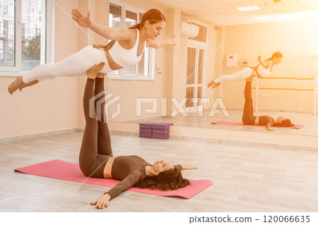Two women do acroyoga on a light floor near a large mirror in a yoga studio. They are wearing leggings and tops. 120066635