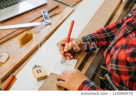 Person in a red plaid shirt sketching table plans on paper with a red pencil, surrounded by wooden planks and miniature houses. Perfect for architecture, construction, and DIY project concepts 120066878