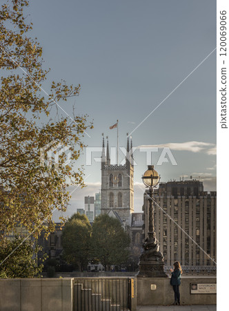 Side view portrait woman standing while VDO call on smart phone next to Lamp post on Thames Path with Southwark Anglican Diocese church tower and Minerva House in the background in the City of London 120069066