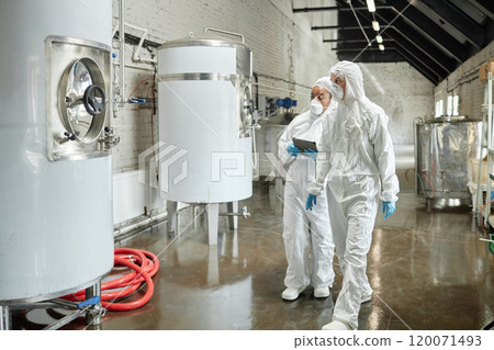 Full length shot of professional female supervisor in mask and coverall walking towards stainless tank with liquid while inspecting workshop equipment with colleague at factory Full length shot of professional female supervisor in mask and coverall walking towards stainless tank with liquid while inspecting workshop equipment with colleague at factory 120071493