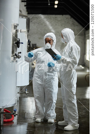 Vertical shot of two professional female supervisors wearing masks and coveralls inspecting tanks in cider factory workshop while collaboratively controlling product production Vertical shot of two professional female supervisors wearing masks and coveralls inspecting tanks in cider factory workshop while collaboratively controlling product production 120071500