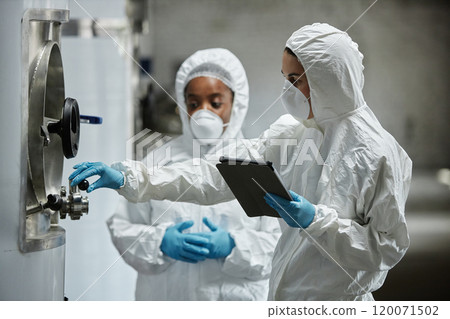 Side view of female process technician in hazmat suit using tablet computer inspecting stainless tanks with colleague while controlling fermentation process at craft cider factory, copy space 120071502
