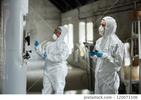 Medium shot of female process technician in hazmat suit using tablet computer inspecting stainless tanks while controlling fermentation process with colleague at craft cider factory, copy space Medium shot of female process technician in hazmat suit using tablet computer inspecting stainless tanks while controlling fermentation process with colleague at craft cider factory, copy space 120071506