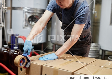 Cropped shot of male production worker packing cardboard boxes with adhesive tape getting kegs with craft beverage for delivering to pubs from factory, copy space 120071622