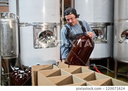 Medium shot of hard working male production operator packing huge keg of craft cider or beer into cardboard box for delivering to pub from factory, copy space 120071624